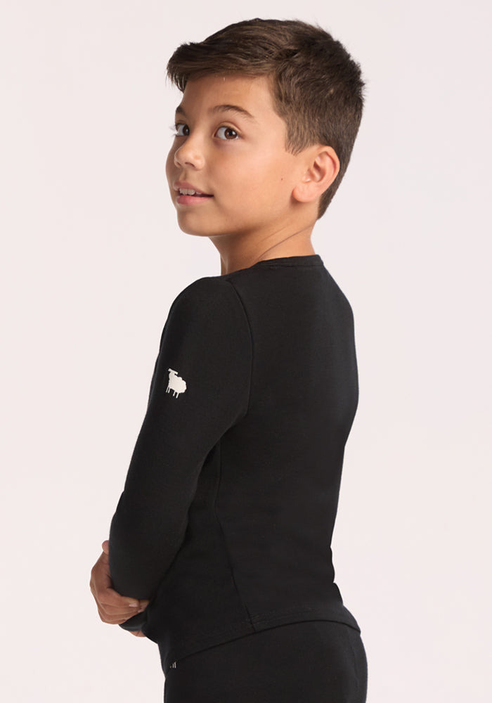 A young boy with short brown hair, arms crossed, looks over his shoulder against a plain light background, wearing the Woolx Kids Explorer Base Layer Top in black with a small white sheep logo on the sleeve.