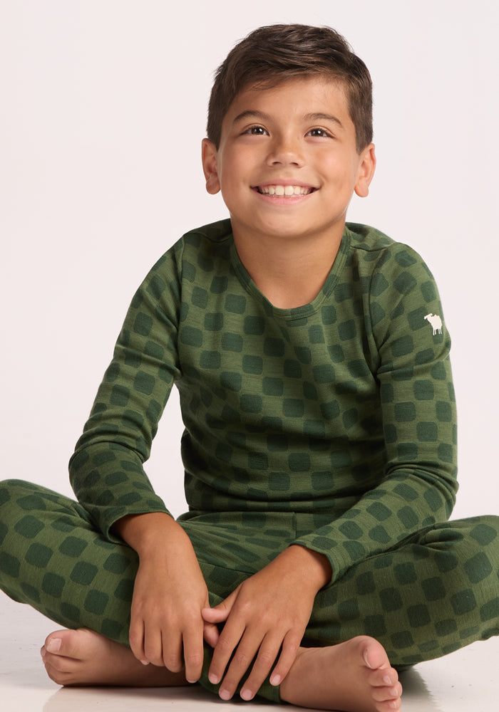 A young boy with short dark hair smiles while sitting cross-legged, wearing the Woolx Kids Explorer Base Layer Top - Frog Hop in green with a dark checkered pattern and a small white animal logo on the sleeve. The background is plain white.