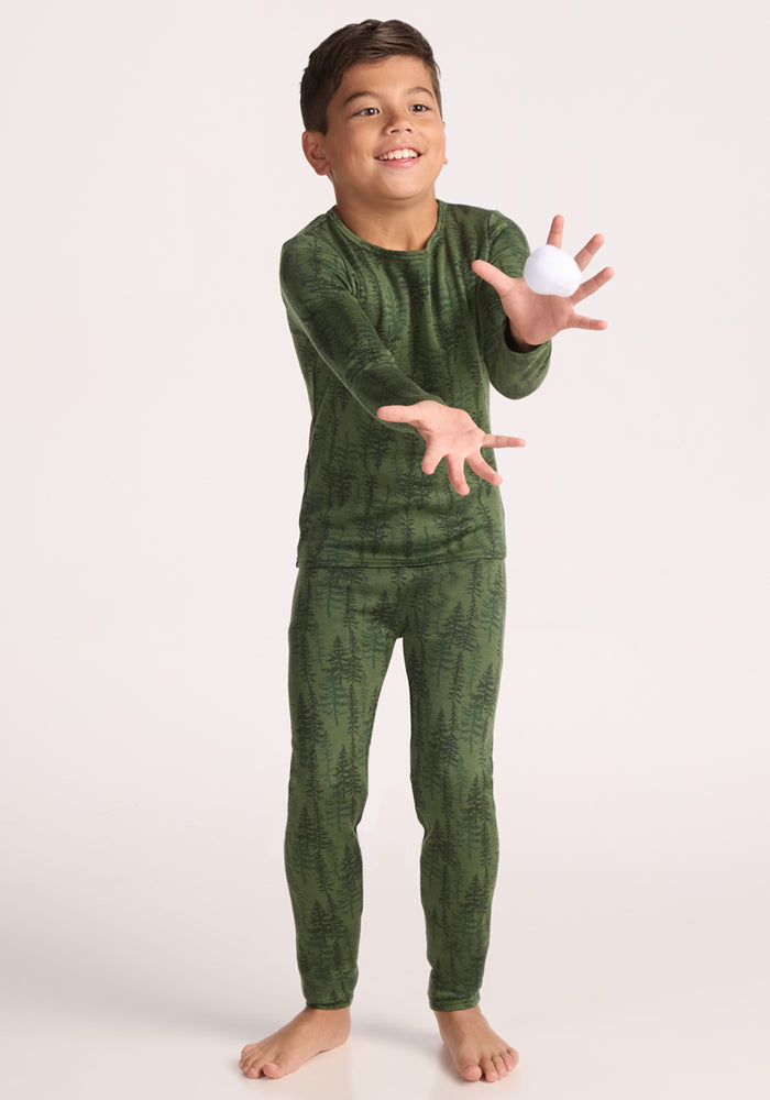 A young boy stands barefoot against a plain light background, reaching out to catch a white ball while wearing Woolx Kids Explorer Base Layer Bottoms - Forest Friends.