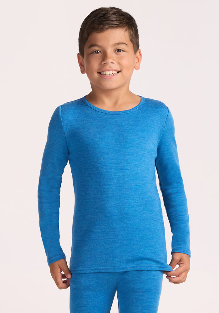 A young boy smiles at the camera wearing the Woolx Kids Explorer Base Layer Top in Blue Razz, made from soft Australian Merino wool. He stands against a plain light background, holding the edge of his shirt with one hand. 
