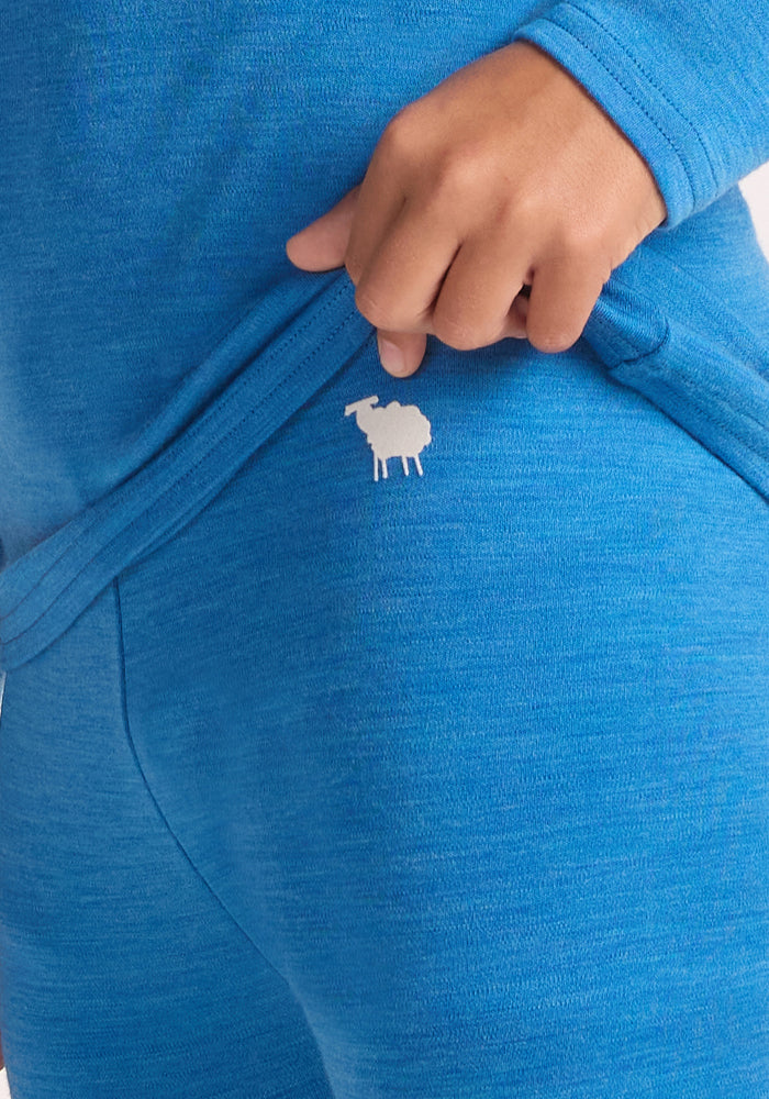 A child in blue raises their shirt to show a small white sheep logo on the waistband of Woolx Kids Explorer Base Layer Bottoms - Blue Razz.