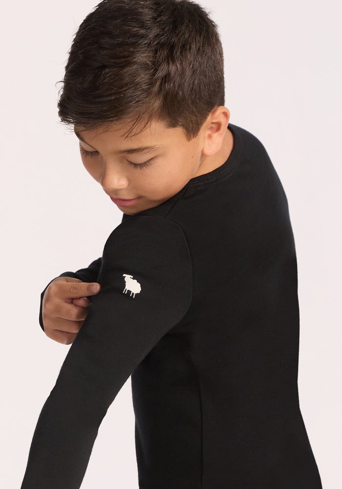 A boy with short brown hair wears the Woolx Kids Explorer Base Layer Top in black and looks over his shoulder at a small white sheep logo on the sleeve. The background is plain and light-colored.