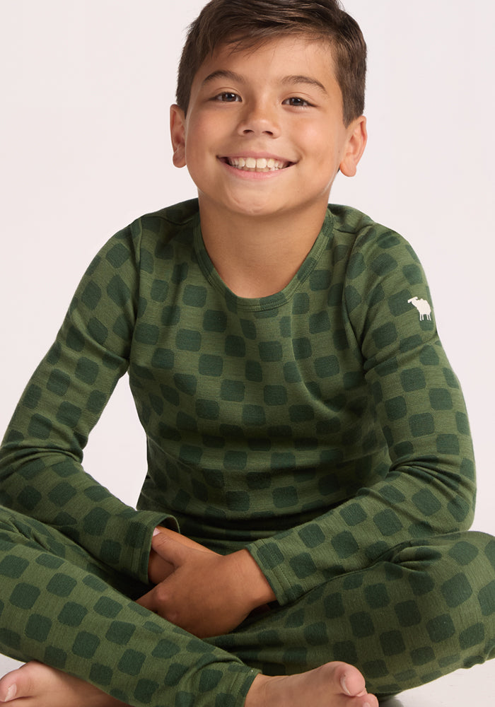 A smiling young boy with short brown hair sits cross-legged in matching green pajamas, wearing Woolx Kids Explorer Base Layer Bottoms - Frog Hop with a dark square pattern and a small white animal logo on the shoulder.