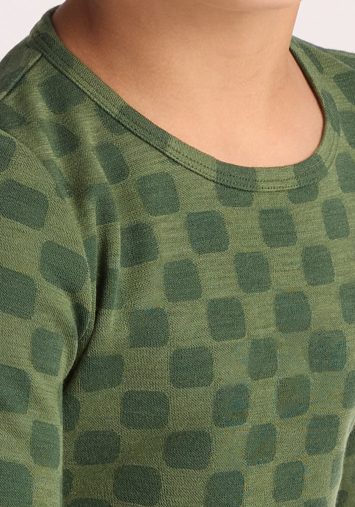 A close-up shows a child wearing the Woolx Kids Explorer Base Layer Top - Frog Hop, featuring green breathable wool with a darker green square pattern. Only the lower face, neck, shoulder, and upper chest are visible.