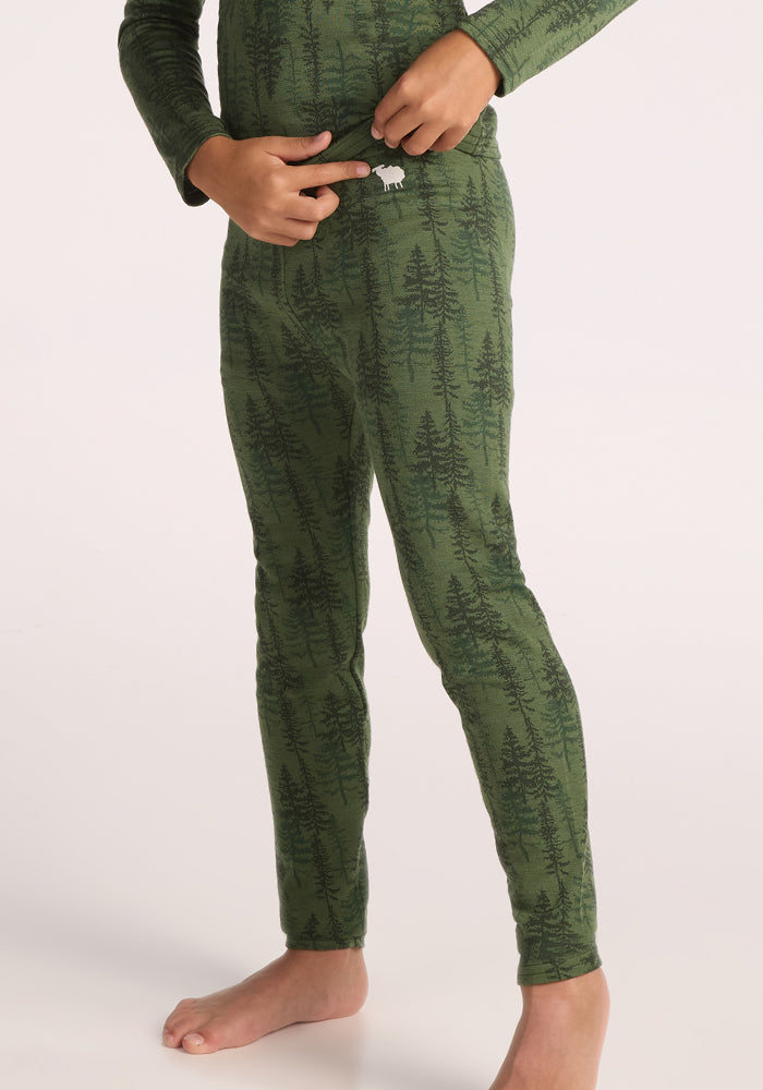 A child in green Kids Explorer Base Layer Bottoms - Forest Friends by Woolx stands barefoot, one hand on the waistband and the other on their hip. Their upper body and face are not shown.