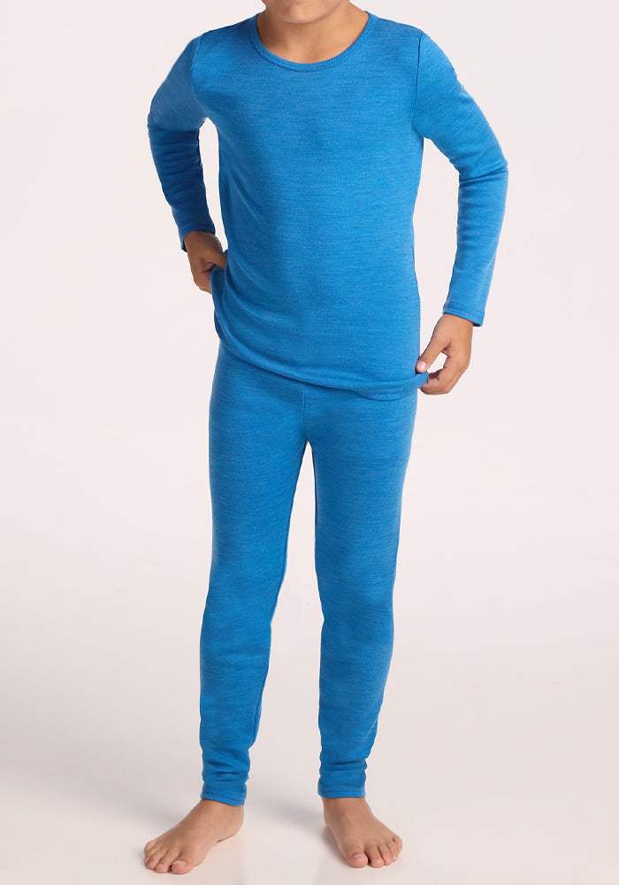 A child stands barefoot against a plain white background, wearing Woolx Kids Explorer Base Layer Bottoms in Blue Razz with a matching long-sleeve top and one hand resting on their hip.