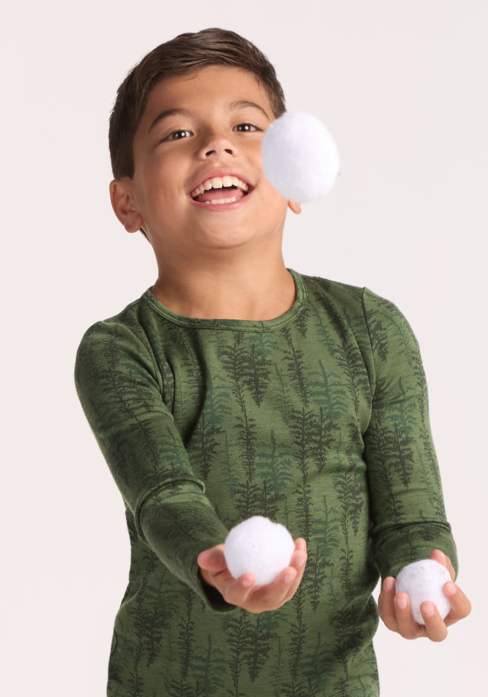 A smiling young boy wears the Woolx Kids Explorer Base Layer Top - Forest Friends, a green merino wool shirt with a playful tree pattern, as he juggles three white balls against a plain background.