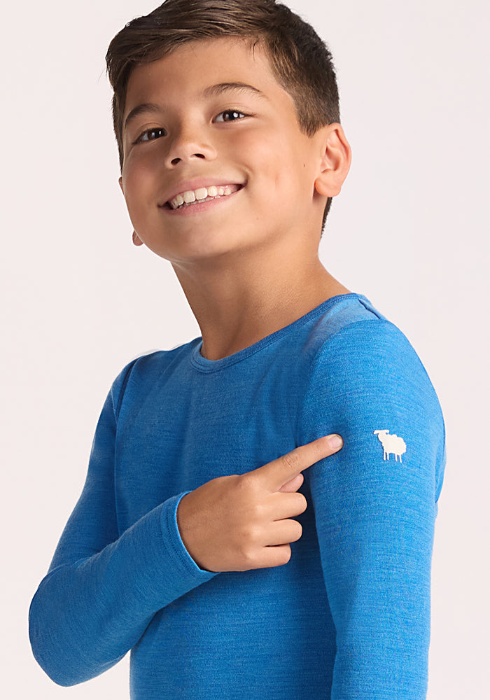 A smiling boy in a Woolx Kids Explorer Base Layer Top - Blue Razz points to a small white sheep logo on his upper arm.