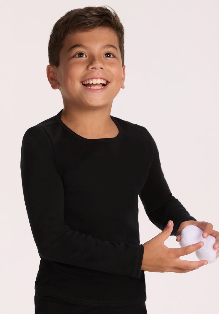 A smiling young boy wears the Woolx Kids Explorer Base Layer Top in black, holding a snowball against a plain light background.