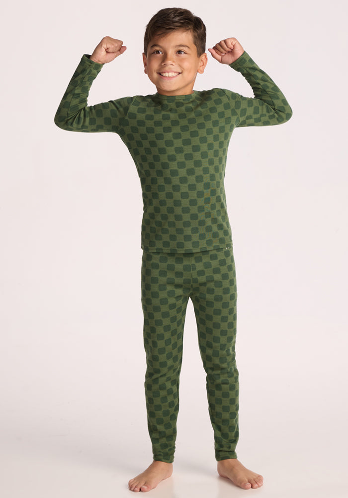 A smiling boy stands barefoot on a white background, wearing the Woolx Kids Explorer Base Layer Top - Frog Hop. He raises both arms and flexes his muscles in a playful pose.