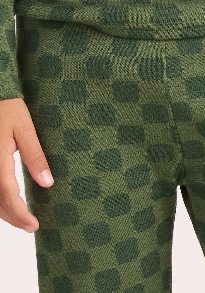 A child's hand rests by their side, wearing the Woolx Kids Explorer Base Layer Bottoms - Frog Hop, featuring green fabric with a darker green checkered pattern. Only the torso, legs, and part of an arm and hand are visible.