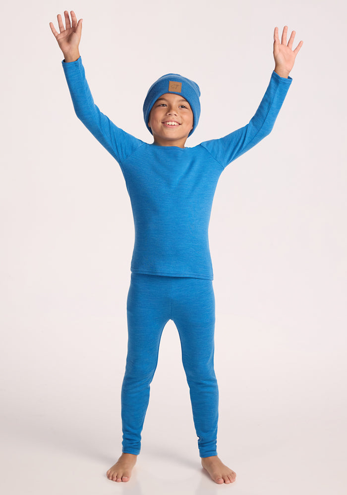 A smiling child stands barefoot with arms raised against a plain background, wearing Woolx Kids Explorer Base Layer Bottoms in Blue Razz, paired with a matching top and beanie.