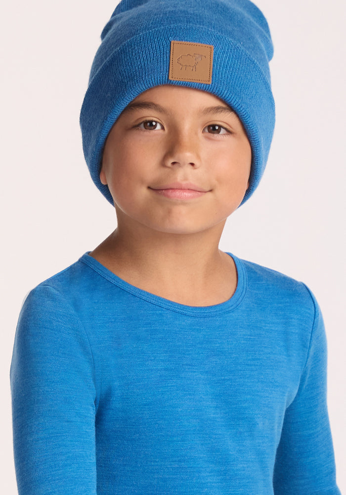 A young child in a Woolx Kids Explorer Base Layer Top in Blue Razz and a matching blue beanie with a brown leather patch stands against a plain light background.