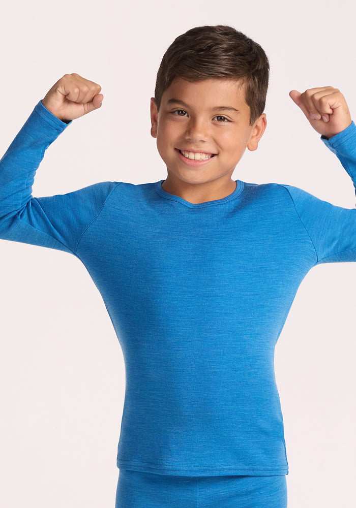 A smiling young boy models the Woolx Kids Explorer Base Layer Top in Blue Razz, flexing his arms confidently against a plain light background.
