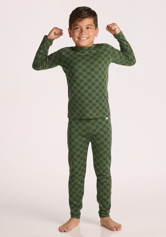A young boy stands barefoot on a white background, smiling and flexing his arms while wearing Woolx Kids Explorer Base Layer Bottoms - Frog Hop in green plaid.