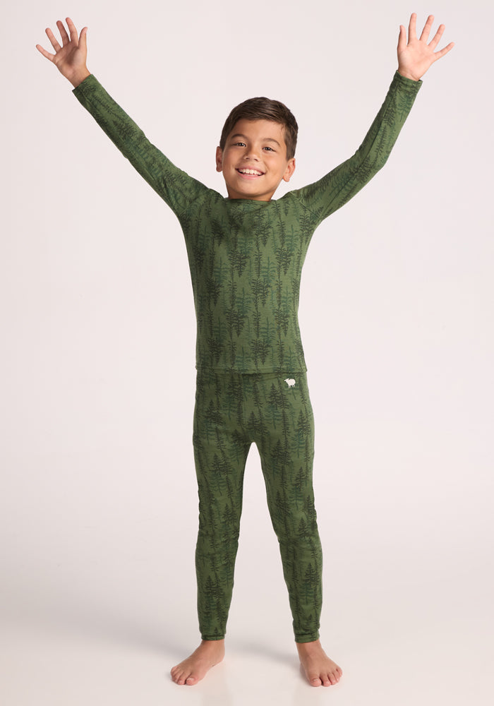 A smiling child stands barefoot with arms raised and eyes closed, wearing the Woolx Kids Explorer Base Layer Top - Forest Friends, against a plain light background.