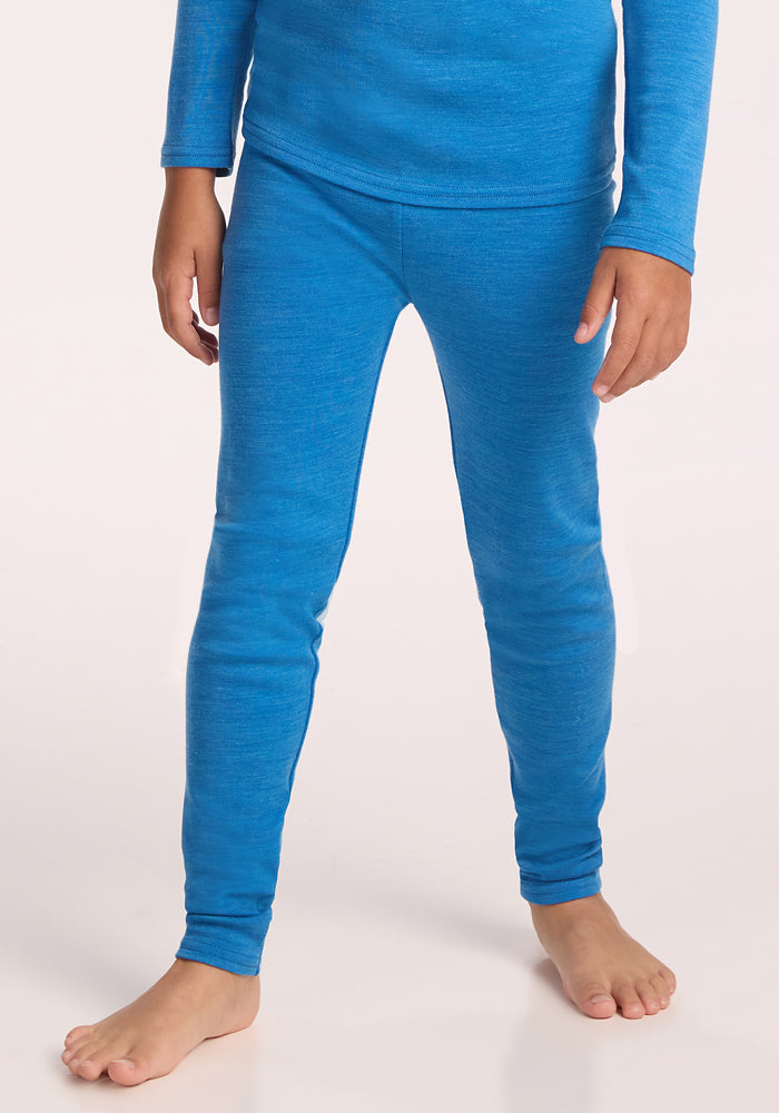 A child stands barefoot against a light background, shown from the neck down, wearing Woolx Kids Explorer Base Layer Bottoms in Blue Razz with a matching long-sleeve top. Only the body, arms, and legs are visible. 
