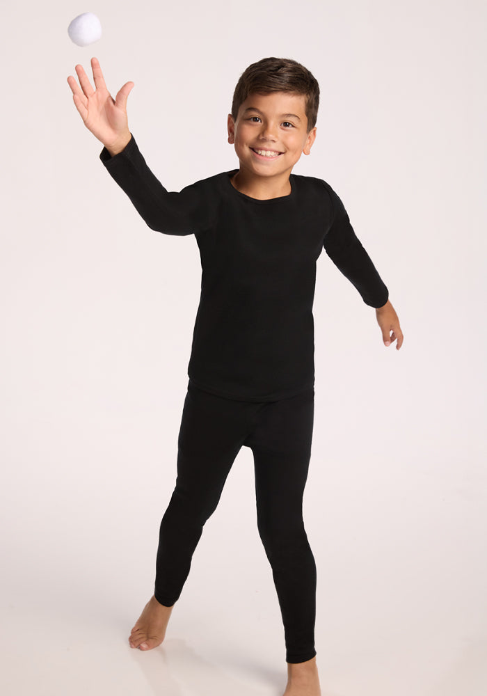 A young boy in a Woolx Kids Explorer Base Layer Top - Black smiles while tossing a white ball with one hand against a plain, light background.