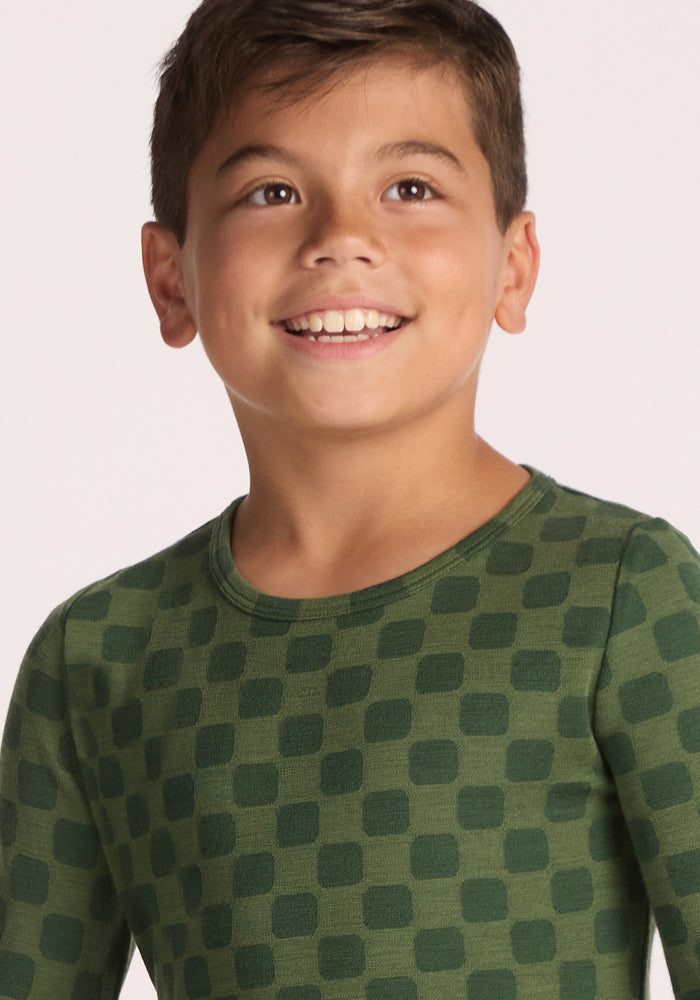 A young boy with short brown hair smiles and looks up, wearing the Woolx Kids Explorer Base Layer Top - Frog Hop, a breathable green wool thermal kids' top with a pattern of darker green squares. The background is plain and light.