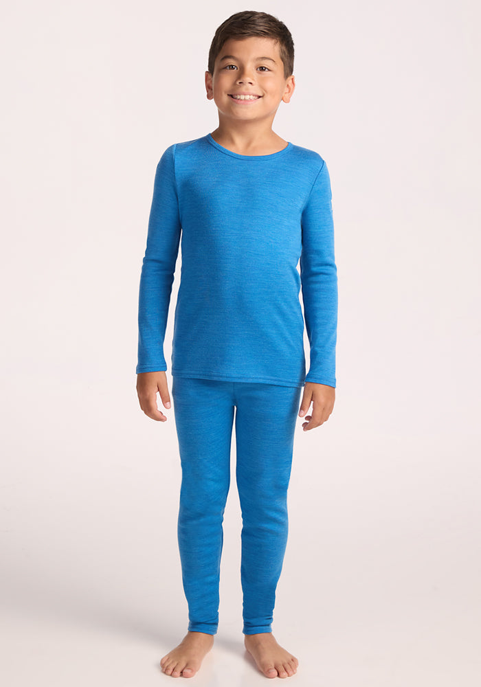 A smiling young boy stands barefoot in a Woolx Kids Explorer Base Layer Top - Blue Razz, paired with matching thermal leggings, against a plain light background.