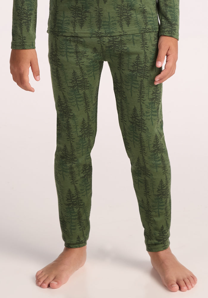 A child stands barefoot in Woolx Kids Explorer Base Layer Bottoms - Forest Friends, cozy green pajama pants with a dark tree pattern, ideal as a warm baselayer or comfy kids ski wear, set against a light background. 