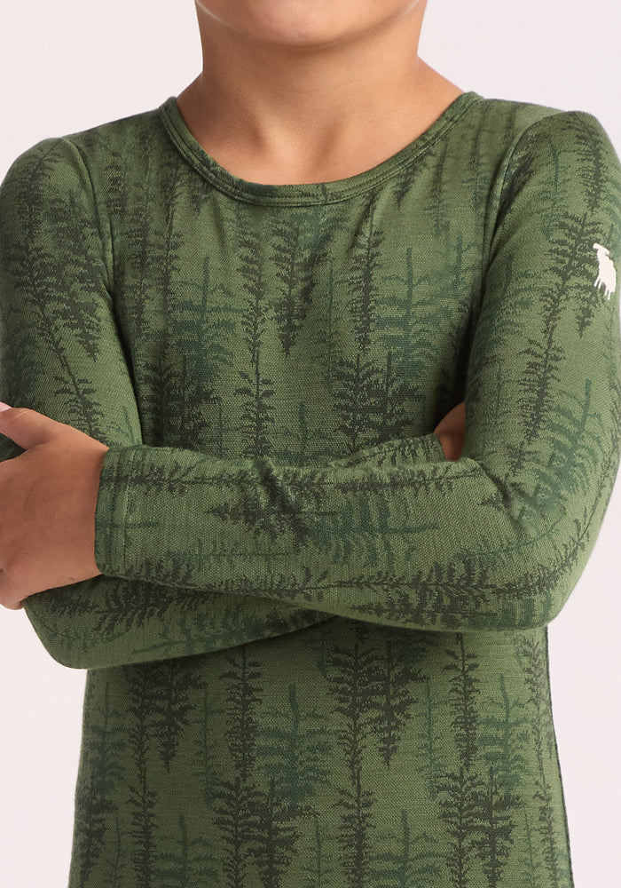 A child with arms crossed wears the Woolx Kids Explorer Base Layer Top - Forest Friends, a green Merino wool shirt featuring a dark fern pattern and a small white moose logo on the shoulder; their face is partially out of frame.