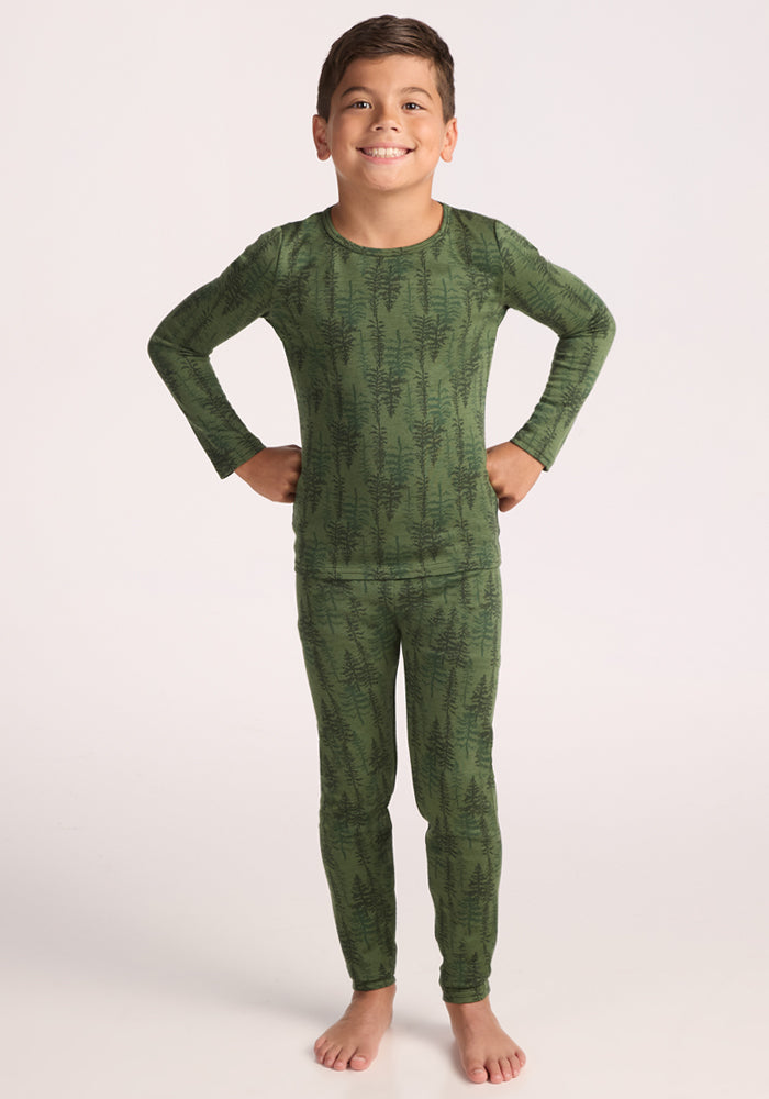 A young child stands barefoot, smiling with hands on hips, wearing Woolx Kids Explorer Base Layer Bottoms - Forest Friends, featuring a matching green top and cozy baselayer bottoms with a dark fern leaf pattern. The background is plain and light-colored.