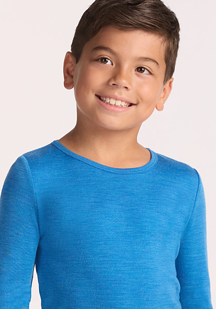 A young boy with short brown hair smiles in a bright blue Woolx Kids Explorer Base Layer Top - Blue Razz, standing against a light background.