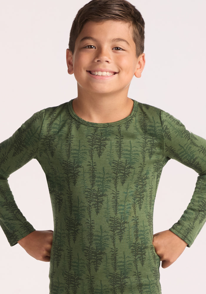 A young boy with short brown hair smiles confidently, wearing the Woolx Kids Explorer Base Layer Top - Forest Friends. He stands with his hands on his hips against a plain light background.
