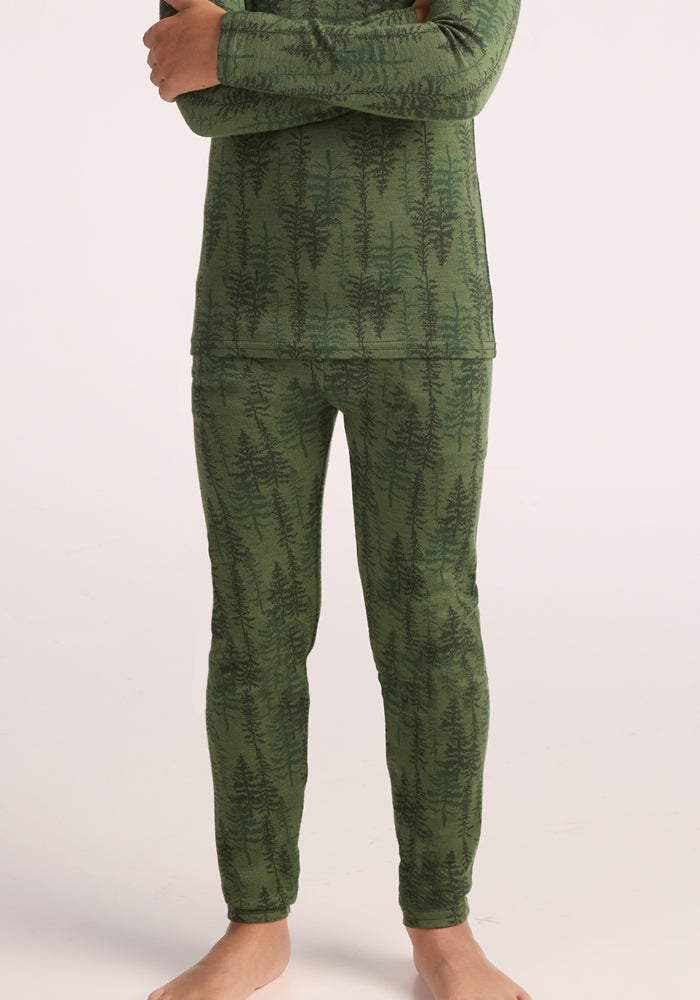 A child models Woolx Kids Explorer Base Layer Bottoms - Forest Friends, featuring a green tree pattern, standing barefoot against a plain light background. The image is cropped at the shoulders.