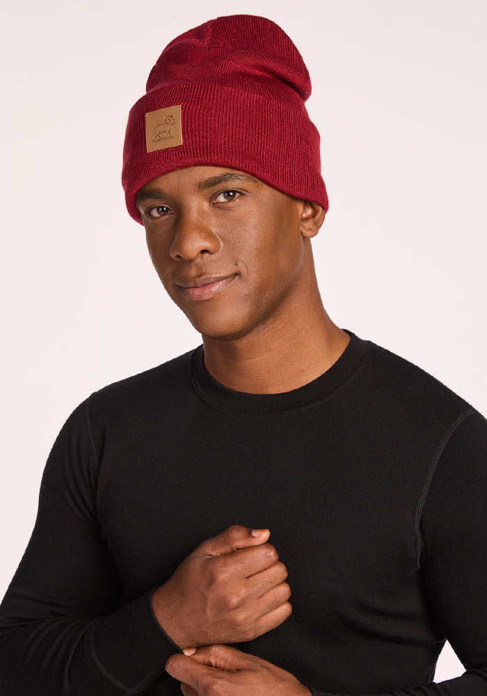 A man in a Woolx Baylor Beanie - Rhubarb, featuring a brown front patch, and a black long-sleeve shirt stands against a light background, looking at the camera with a slight smile.