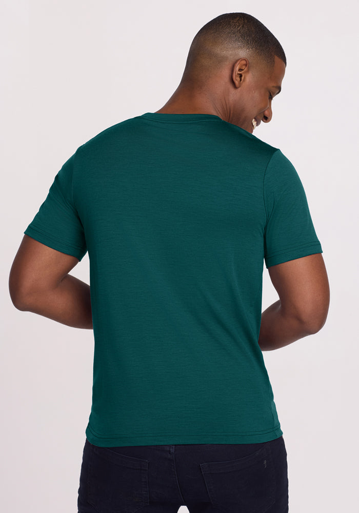 A man faces away from the camera, smiling over his shoulder in a fitted Woolx Endurance Tee - Riverbank Green and black pants, set against a plain light background.