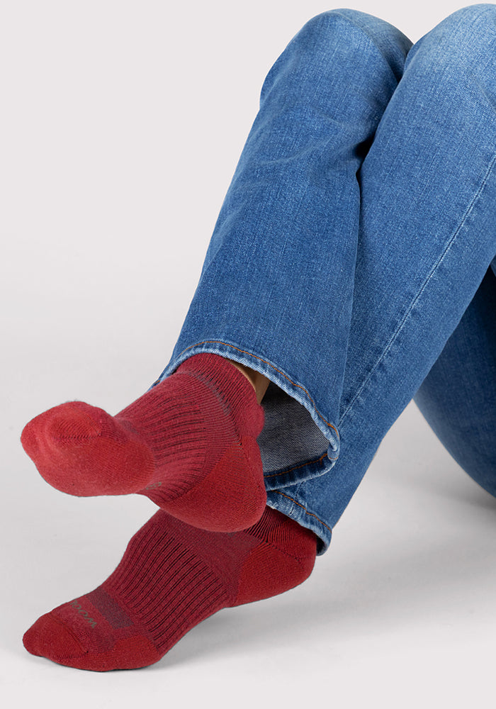 A person wearing blue jeans sits cross-legged, showing their feet in bright rhubarb-colored Womens Rowan No Show Socks by Woolx against a light background.