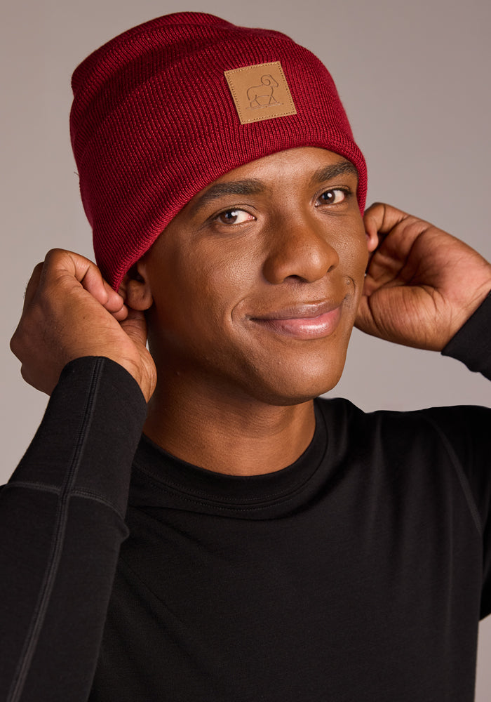 A person in a black long-sleeve shirt smiles while adjusting a Woolx Baylor Beanie - Rhubarb, featuring a tan patch. The background is plain and neutral.