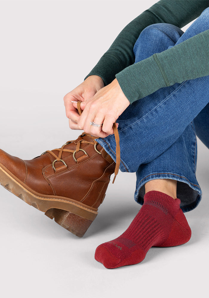 A woman in blue jeans and a green long-sleeve top sits on the floor, tying her brown boot while wearing a Woolx Womens Rowan No Show Sock in Rhubarb on her other foot for comfort and moisture-wicking.