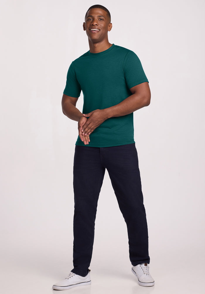 A man stands against a light background, smiling and clasping his hands while wearing the Woolx Endurance Tee in Riverbank Green, paired with dark pants and white sneakers.