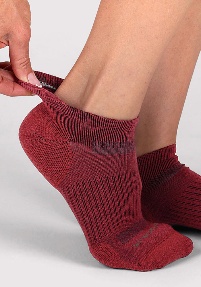 Someone wearing Woolx Womens Rowan No Show Socks in Rhubarb stretches the back of one sock away from their heel. The background is plain light gray.