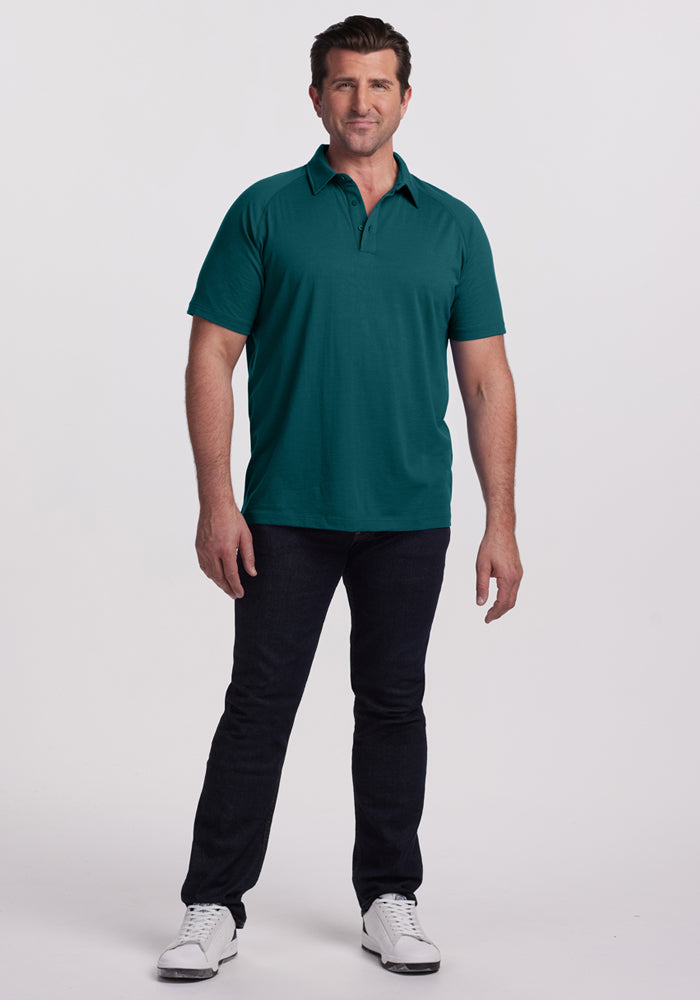 A man faces forward against a white background, wearing the Woolx Summit Polo in Riverbank Green—crafted from merino wool for excellent moisture management—paired with dark jeans and white sneakers.