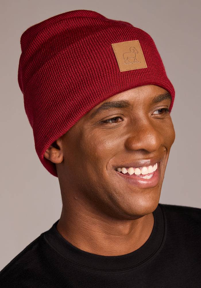 A smiling person wears the Woolx Baylor Beanie - Rhubarb with a tan patch on the front and a black shirt, posed against a plain background.