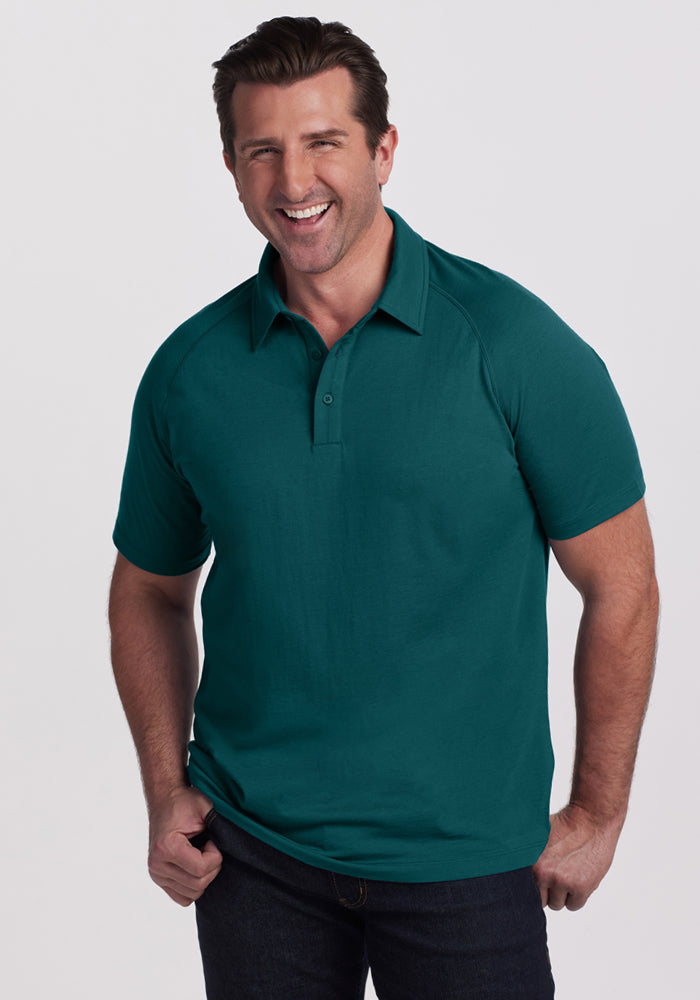 A smiling man with short brown hair wears the Woolx Summit Polo in Riverbank Green and dark jeans. He stands against a plain light background with one hand in his pocket, highlighting the polo’s superior moisture management. 
