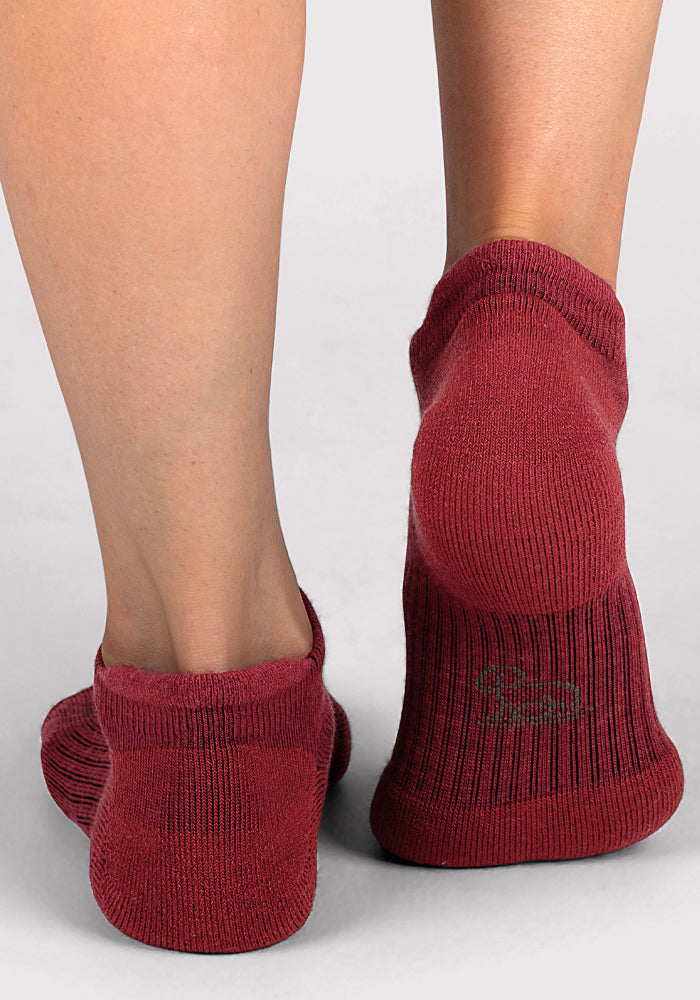 A close-up shows two feet wearing Woolx Womens Rowan No Show Socks in Rhubarb, standing on a light gray surface. The ribbed Merino wool texture is visible, with soft sock lines below the exposed skin above the ankle.
