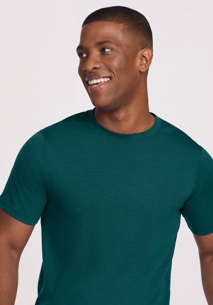 A man with short hair smiles and looks to the side while wearing Woolx’s Endurance Tee in Riverbank Green against a plain light background.