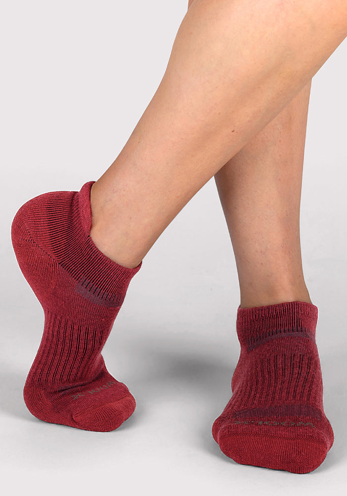 A person stands on tiptoe against a plain, light background, displaying both the side and front view of Woolx Womens Rowan No Show Socks in Rhubarb.