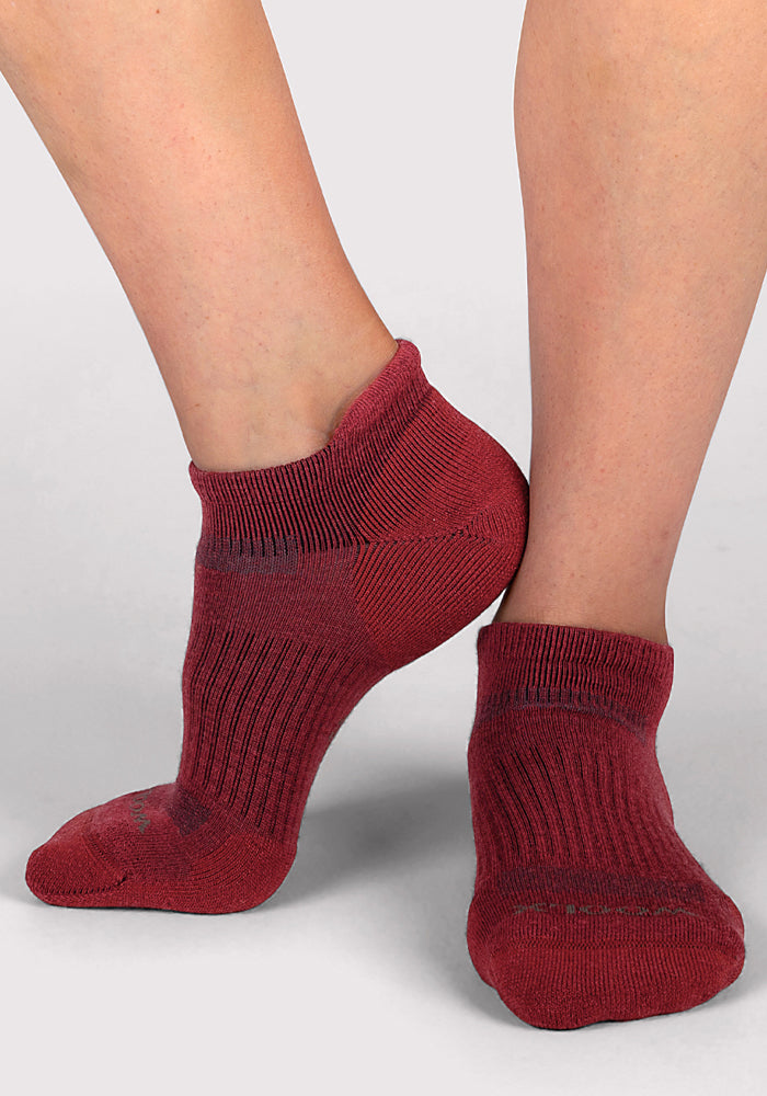 A person stands on tiptoes against a plain light gray background, wearing Woolx Womens Rowan No Show Socks in Rhubarb—a short, maroon Merino wool style with a ribbed texture.