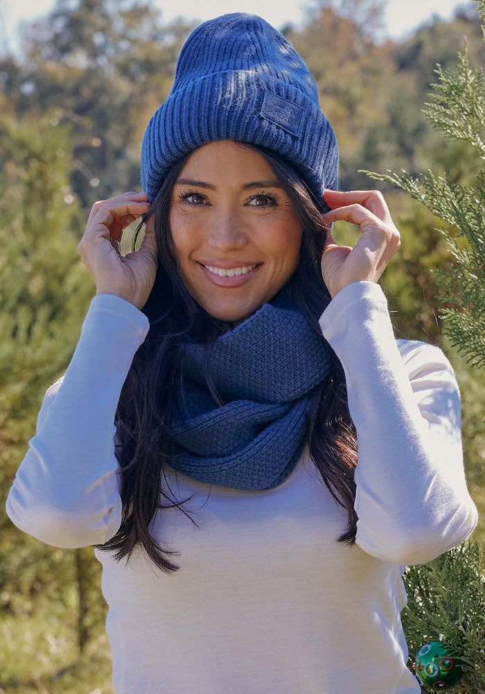 A woman in a blue knit hat and a Woolx Noelle Infinity Scarf - Starry Night smiles outdoors among green trees, wearing a light long-sleeve shirt on a sunny day.