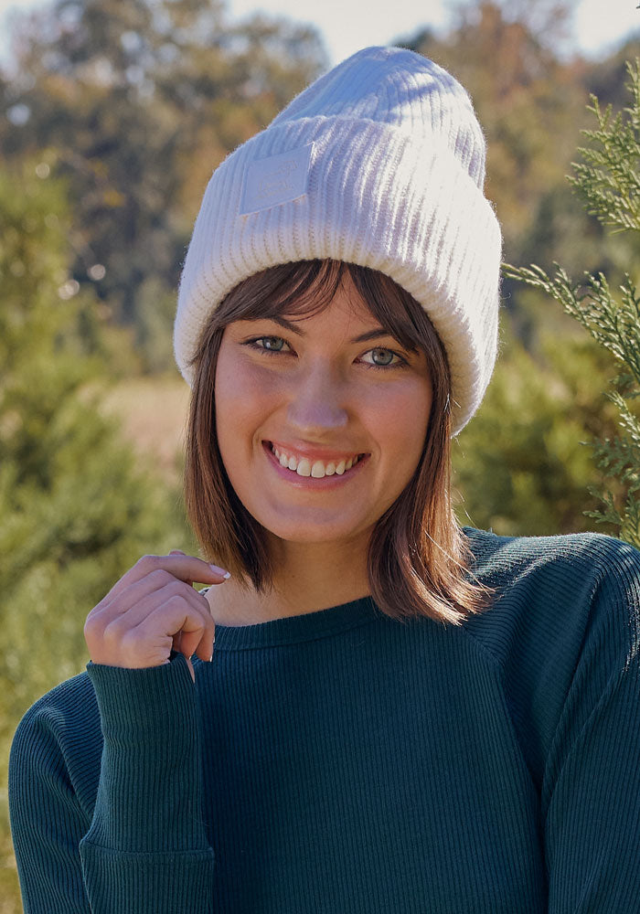A young woman smiles outdoors in soft sunlight, surrounded by green trees, wearing the Woolx Ria Chunky Beanie in Buttercream and a green sweater.