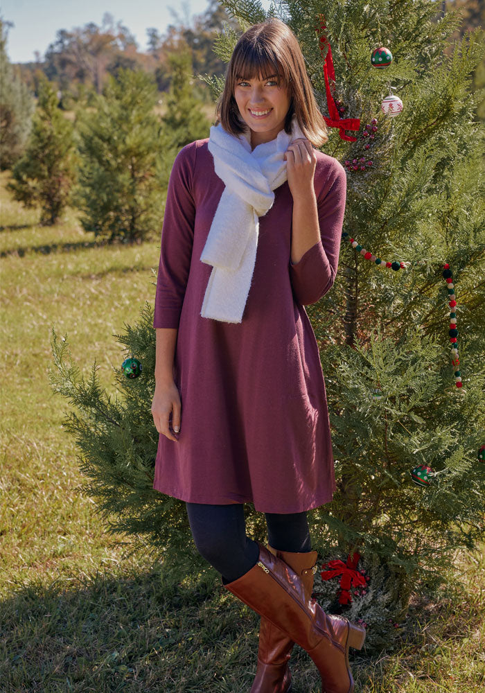 A woman wearing a maroon dress, black leggings, brown boots, and the Woolx Bradie Scarf in Cream smiles beside a decorated outdoor Christmas tree in a grassy area surrounded by evergreens.