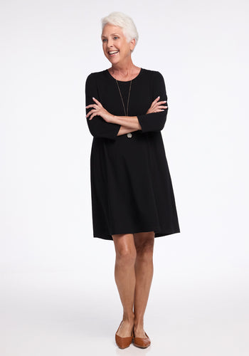 A person with short white hair smiles and stands with crossed arms, wearing the sleek black Rissa A Line Dress by Woolx and brown flat shoes. The plain white background highlights the outfit's travel-friendly simplicity. | Kathy is 5’9”, wearing a size S