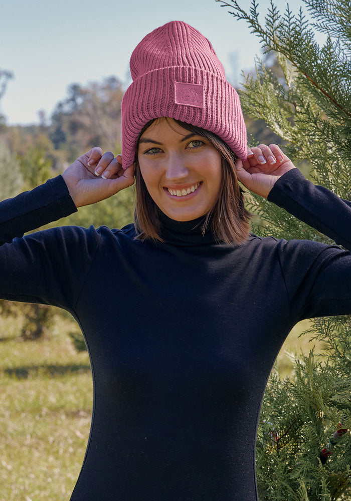 Woman in a Woolx Ria Chunky Beanie in Toasted Rose and black turtleneck smiles outdoors on a sunny day.