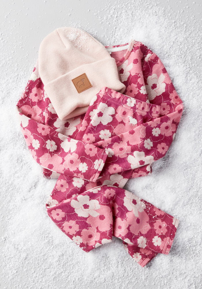 The Woolx Kids Explorer Base Layer Top in Rosie Posie, a pink floral long-sleeve, is displayed on snow beside a light pink knit beanie—highlighting cozy, breathable winterwear for children.